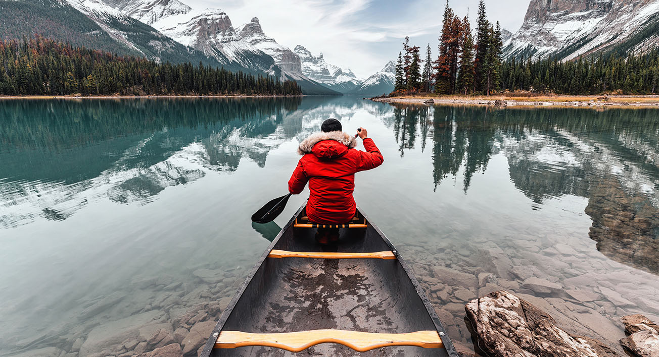 Person paddles a canoe on a lake surrounded by mountains.
