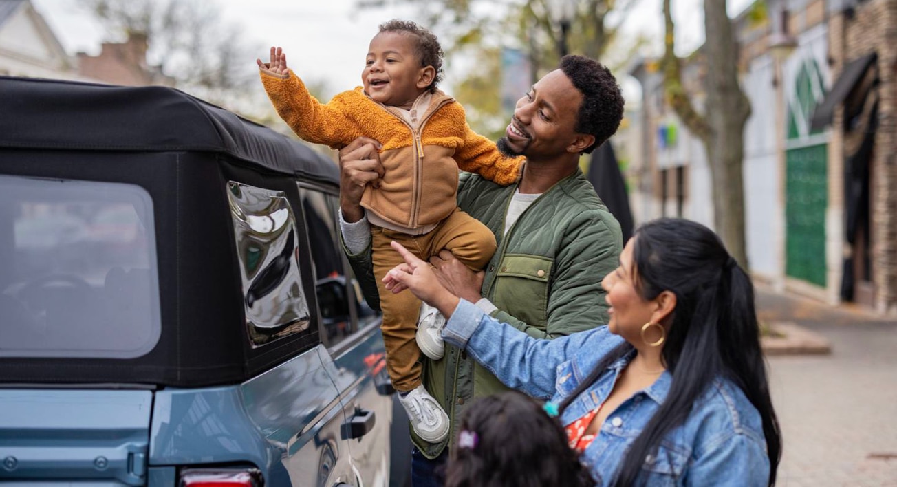 A father lifts his child while the mother smiles and points in front of a Ford Bronco parked on a street.