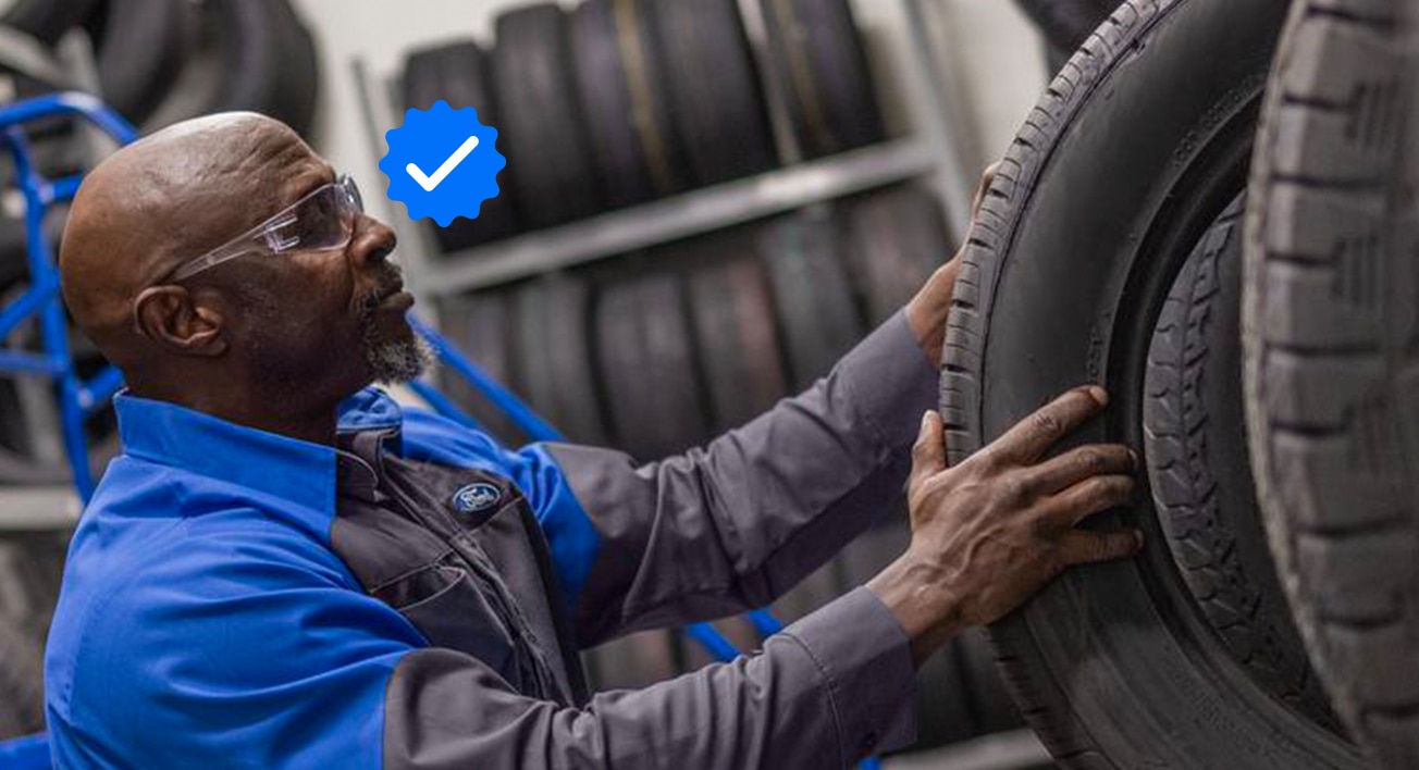 Technicien Service Ford remplaçant un pneu sur un véhicule Ford. Un badge vérifié est visible en surimposition près de la tête du technicien. 