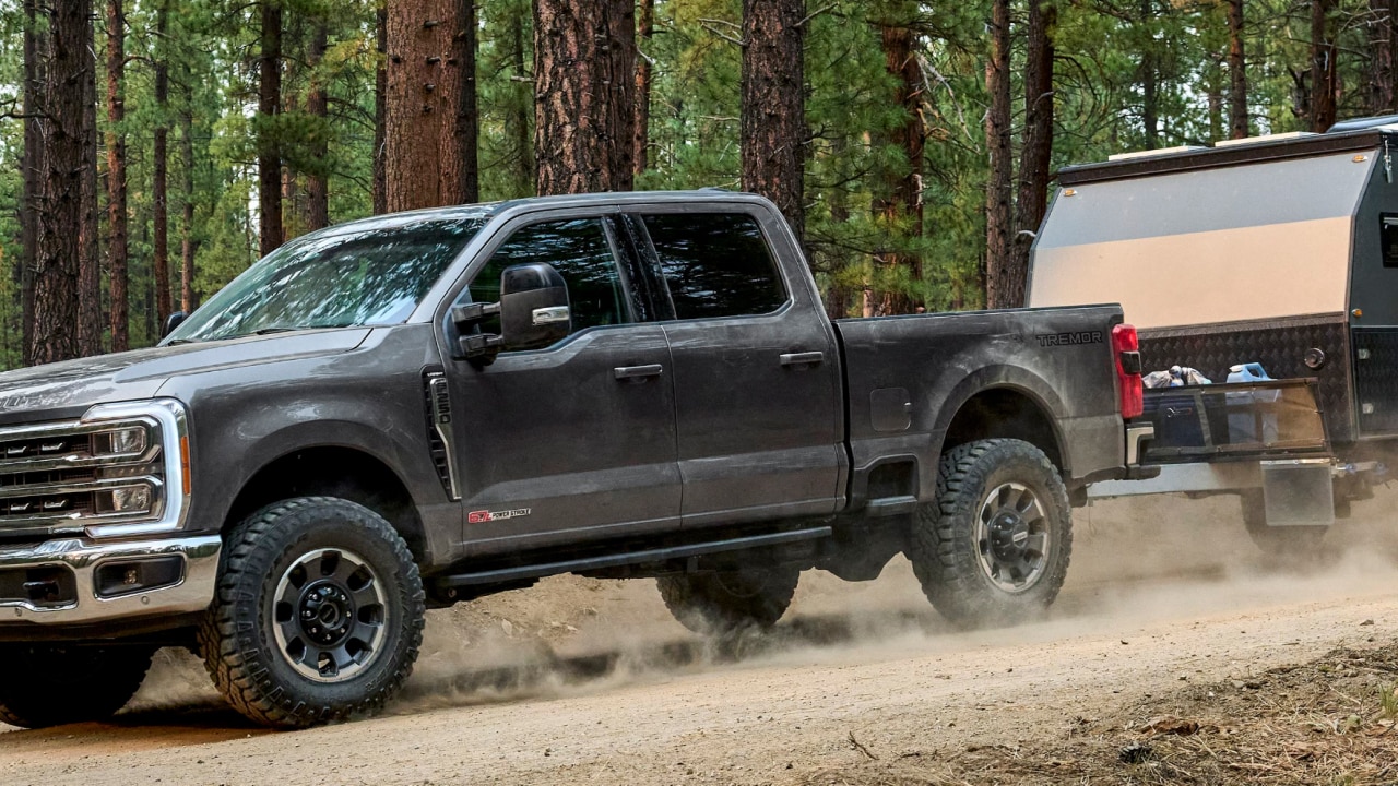 Ford truck towing a camper trailer on a dirt road through a forest.