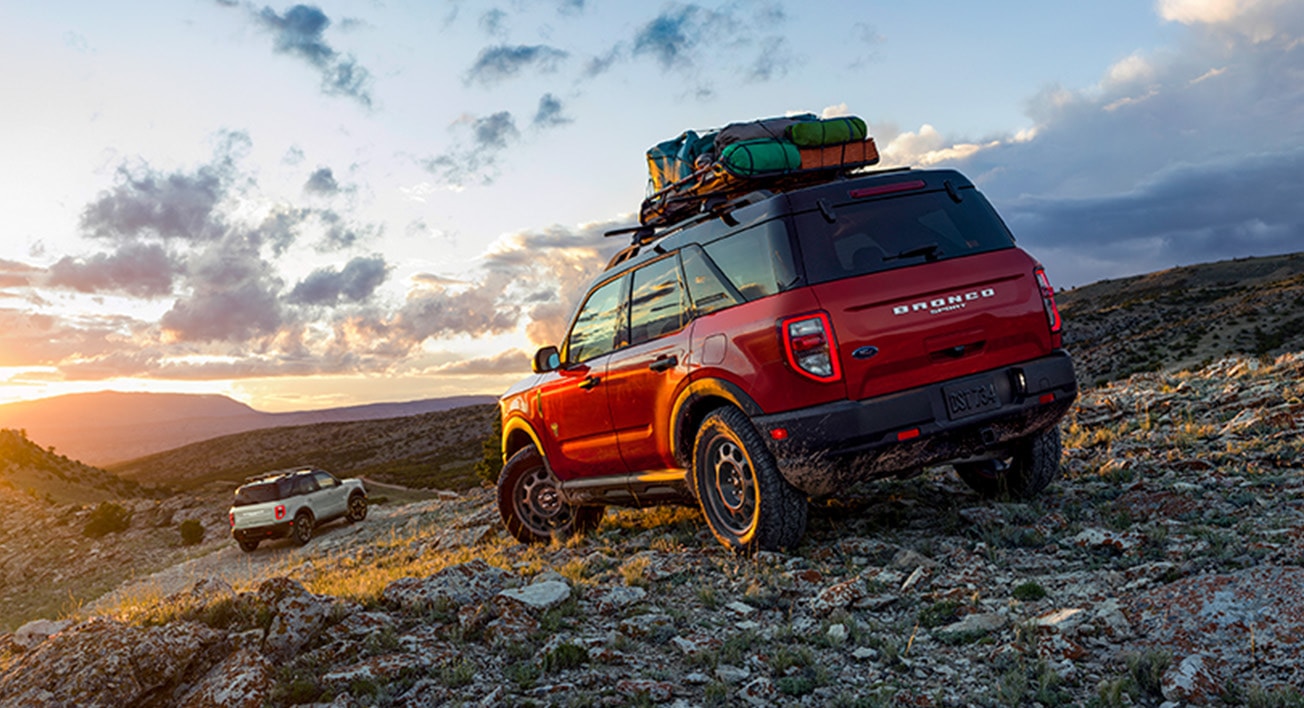 Two Ford Broncos on a rocky trail at sunset, one with roof gear, overlooking a scenic mountain view