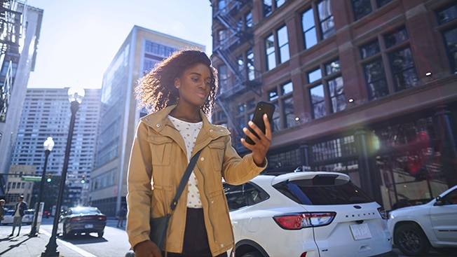 A young woman walking on a sunny city street while looking at her phone.