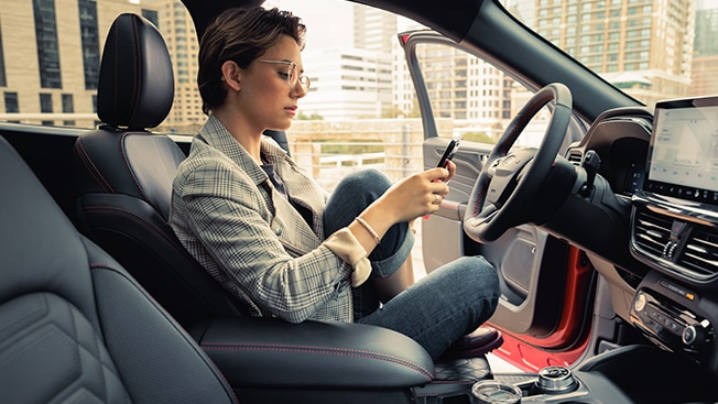A woman sitting inside a Ford Escape SUV looks at her iPhone with the car door open.