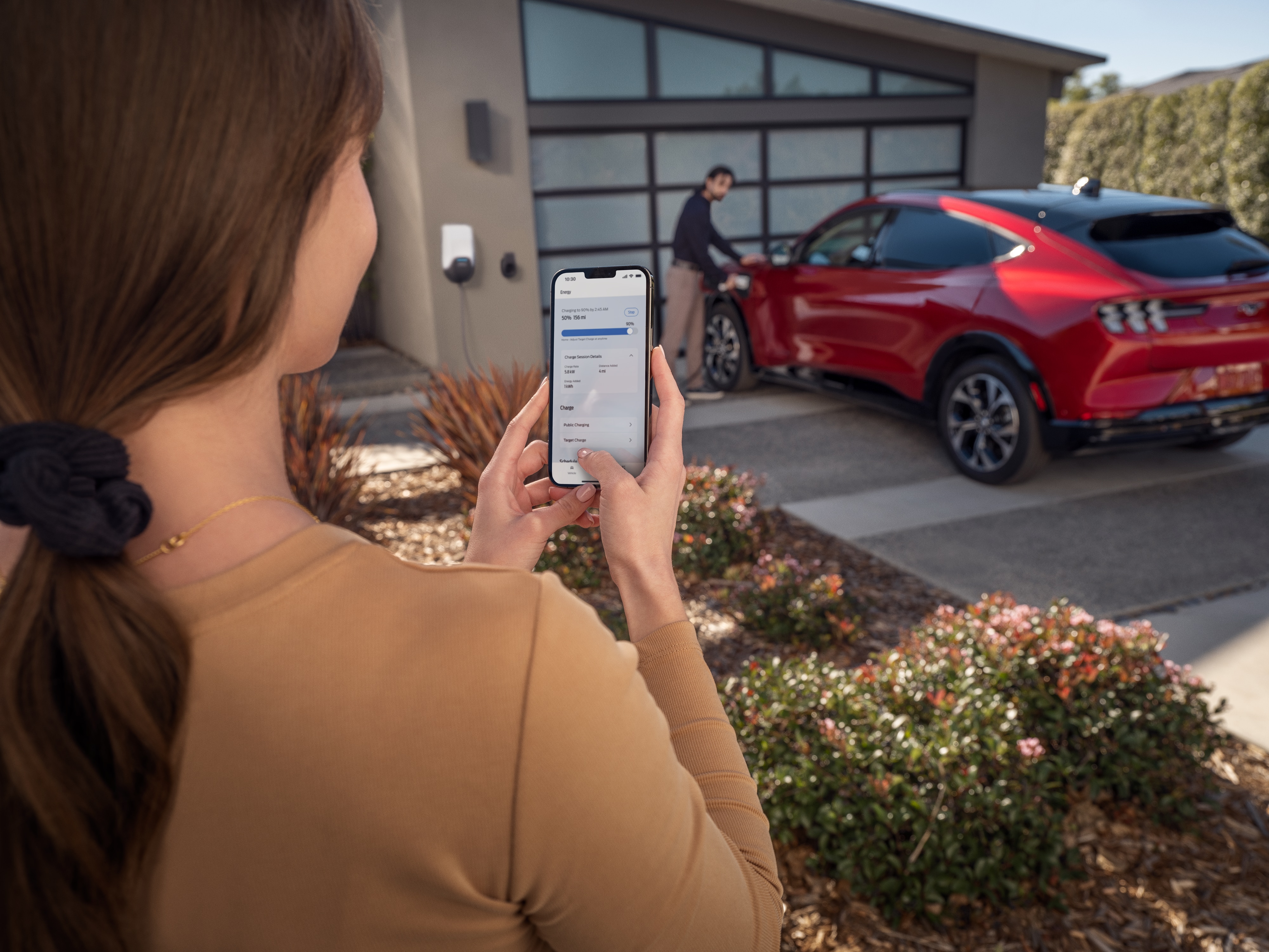 A woman looks at the FordPass App on her iPhone while a man charges his Ford Mustang Mach-E SUV in the background.