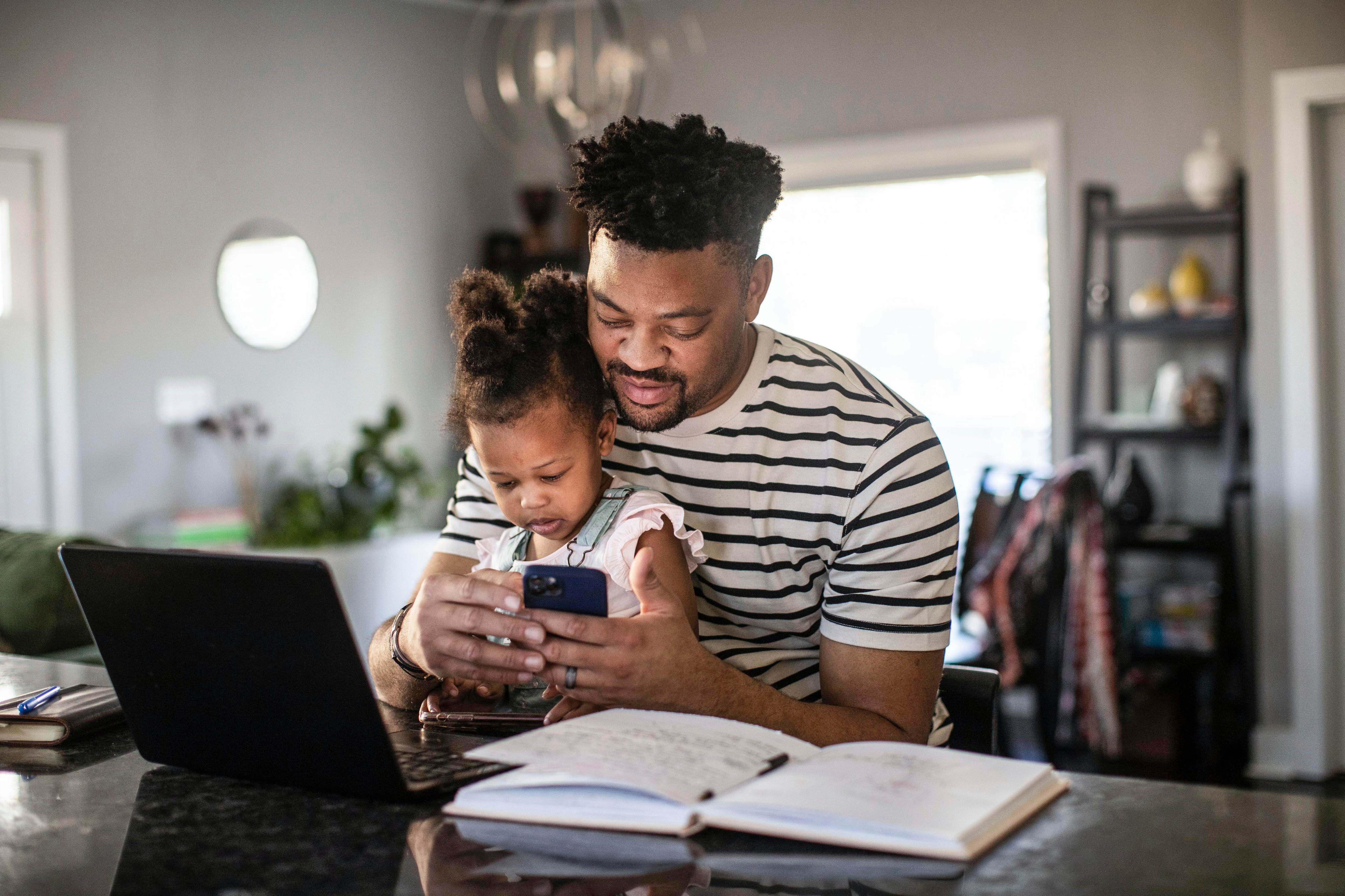 A man checking his phone at home while sitting with his young daughter.