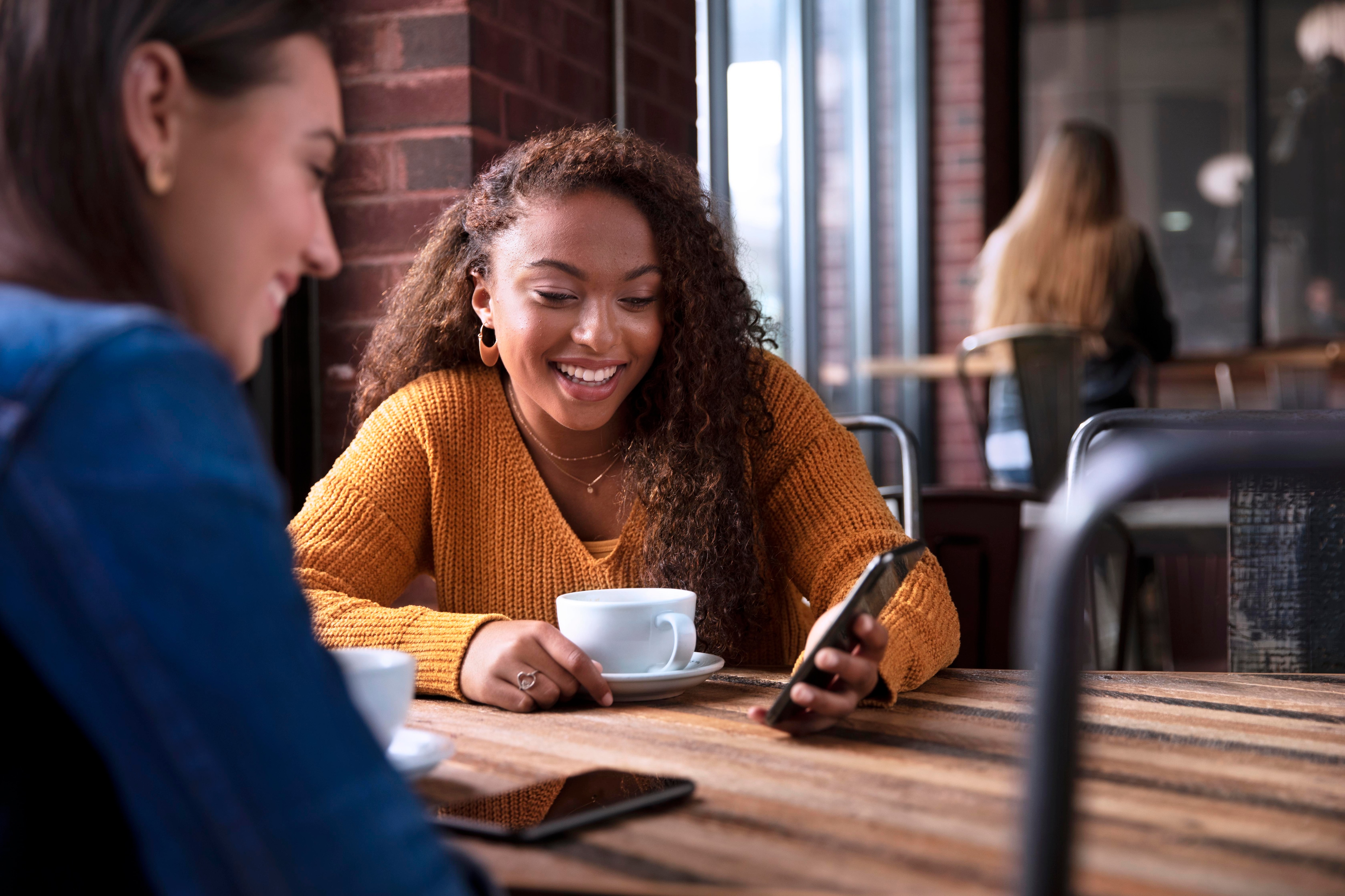 A young woman checking her phone while sitting in a coffeehouse with a friend.