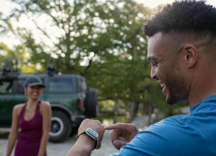 A man interacts with his Apple Watch. A woman is in the background with a Ford Bronco SUV.