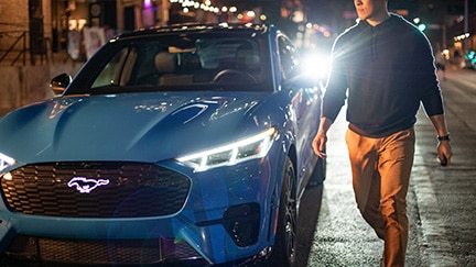 A man walks around the front of a Ford Mustang Mach-E SUV at night.
