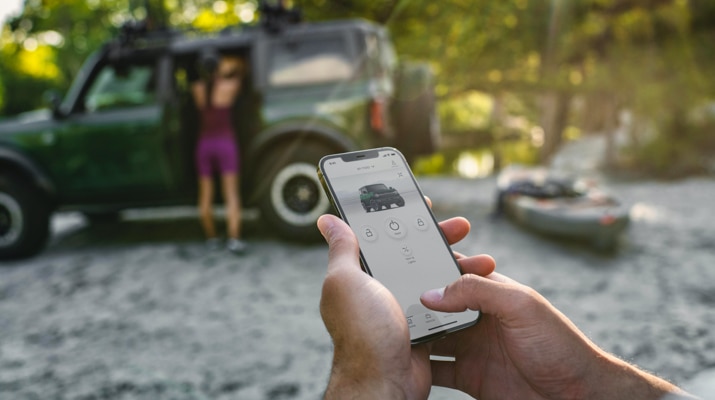 Una persona sostiene un iPhone con la Aplicación FordPass en la pantalla y se ve una SUV Ford Bronco está estacionada de fondo.