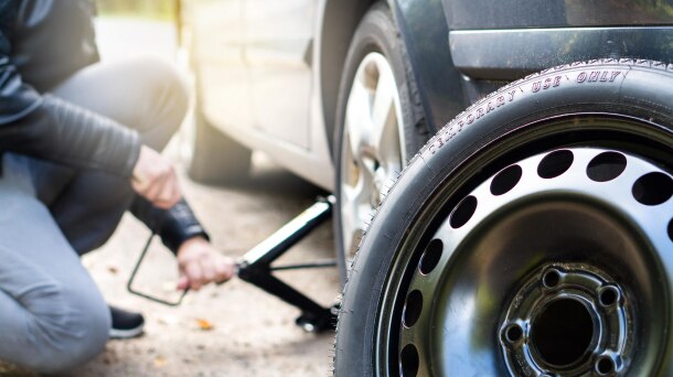 Person kneeling beside vehicle and spare tire holding a car jack.