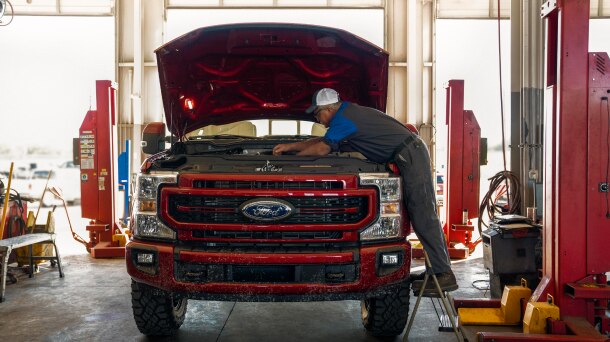Technician standing on stepstool looking under hood of red Ford pickup.