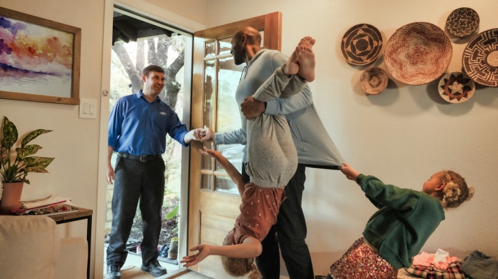 A man opens the door to his home to greet a Ford Service technician. He hands the tech his keys while his two children hang from him and tug on his clothes.