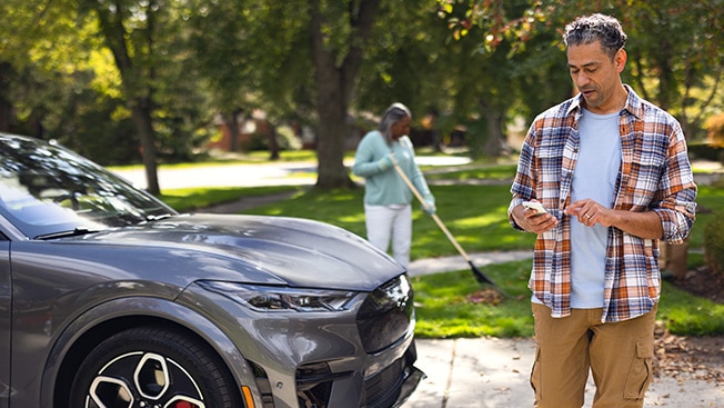 A Ford owner stands in the driveway in front of a parked Mustang Mach-E while using the FordPass App.