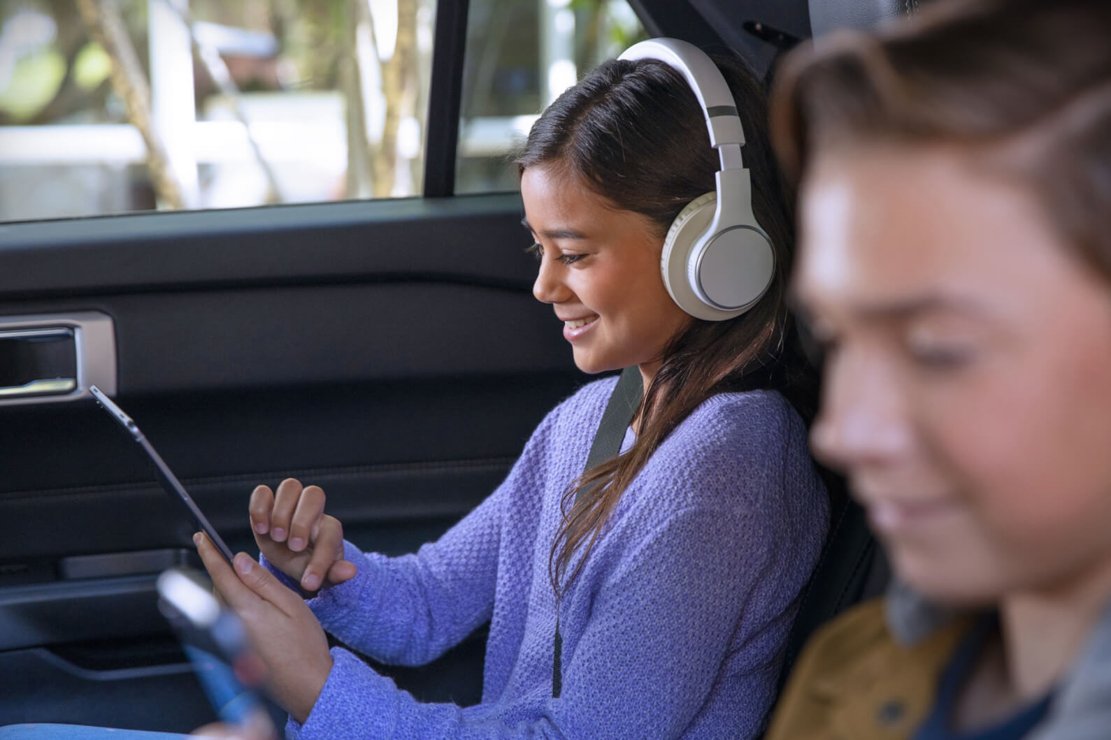 Dos niños con cinturones de seguridad en el asiento trasero de un vehículo Ford mirando sus dispositivos móviles y escuchando con sus auriculares.