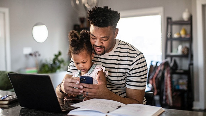 An adult and their child are seated at a desk looking at a phone, with a laptop and notebook open in front of them.