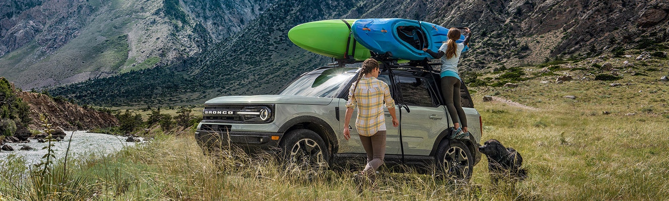 An owner, joined by a friend and their dog, unstraps a kayak from the roof rack of the Bronco Sport® SUV parked in the tall grass beside a glacial lake.