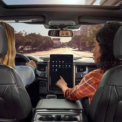 A passenger interacts with the center screen of a Ford vehicle while their friend drives down a tree-lined city street.