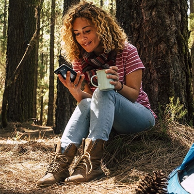 A Bronco owner checks their FordPass® app while leaning against an old-growth pine.