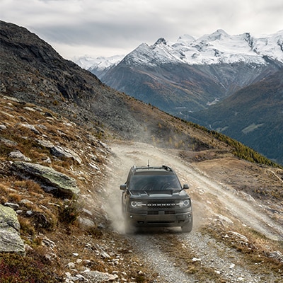 The Bronco Sport® SUV kicks up dust on an elevated trail with snow-topped mountains in the background.