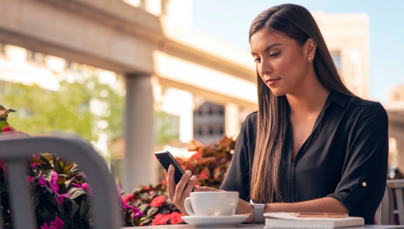 Una mujer sentada a una mesa de café al aire libre sostiene su smartphone y observa la aplicación Lincoln Way.