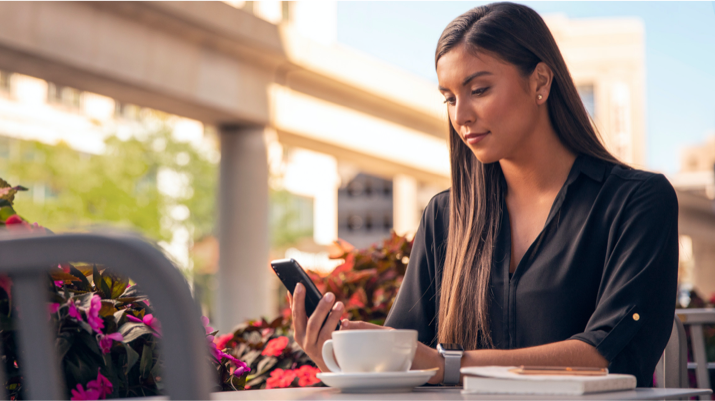 Una mujer sentada a una mesa de café al aire libre sostiene su smartphone y observa la aplicación Lincoln Way.