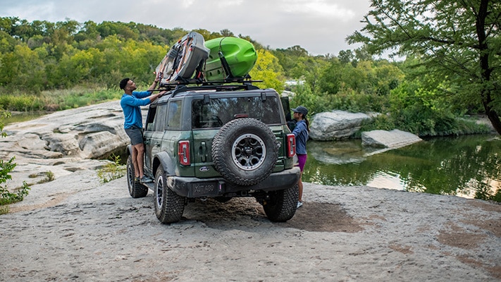 Two drivers standing outside their Bronco® SUV on a riverbank while tying two kayaks to the available roof rack accessory.