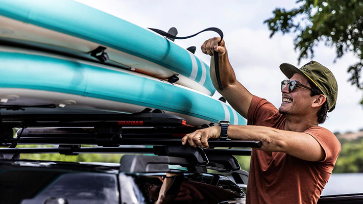 A driver smiling while standing outside their Ford vehicle and tying two paddle boards to the available roof rack accessory.