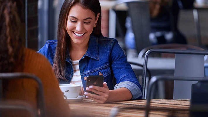 A young woman sitting in a coffeehouse with friends while also looking at her mobile device.