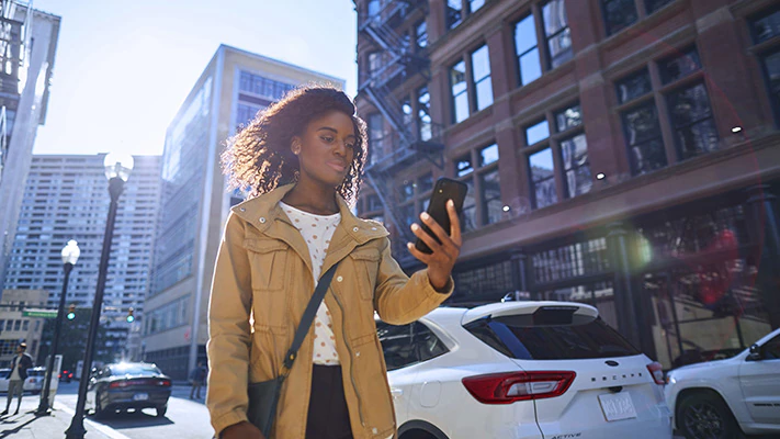 A driver walking down a city street holding a smartphone with a white 2023 Ford Escape® SUV parked nearby.